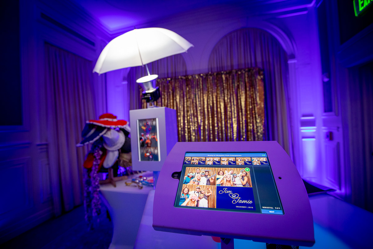 A photobooth setup in a dimly lit room with blue lighting offers a unique touch for events, courtesy of our Photo Booth Rental Services. A tablet displays photo previews, while hats and glasses adorn a table. The backdrop features golden sequins, enhanced by a large umbrella light in the background.